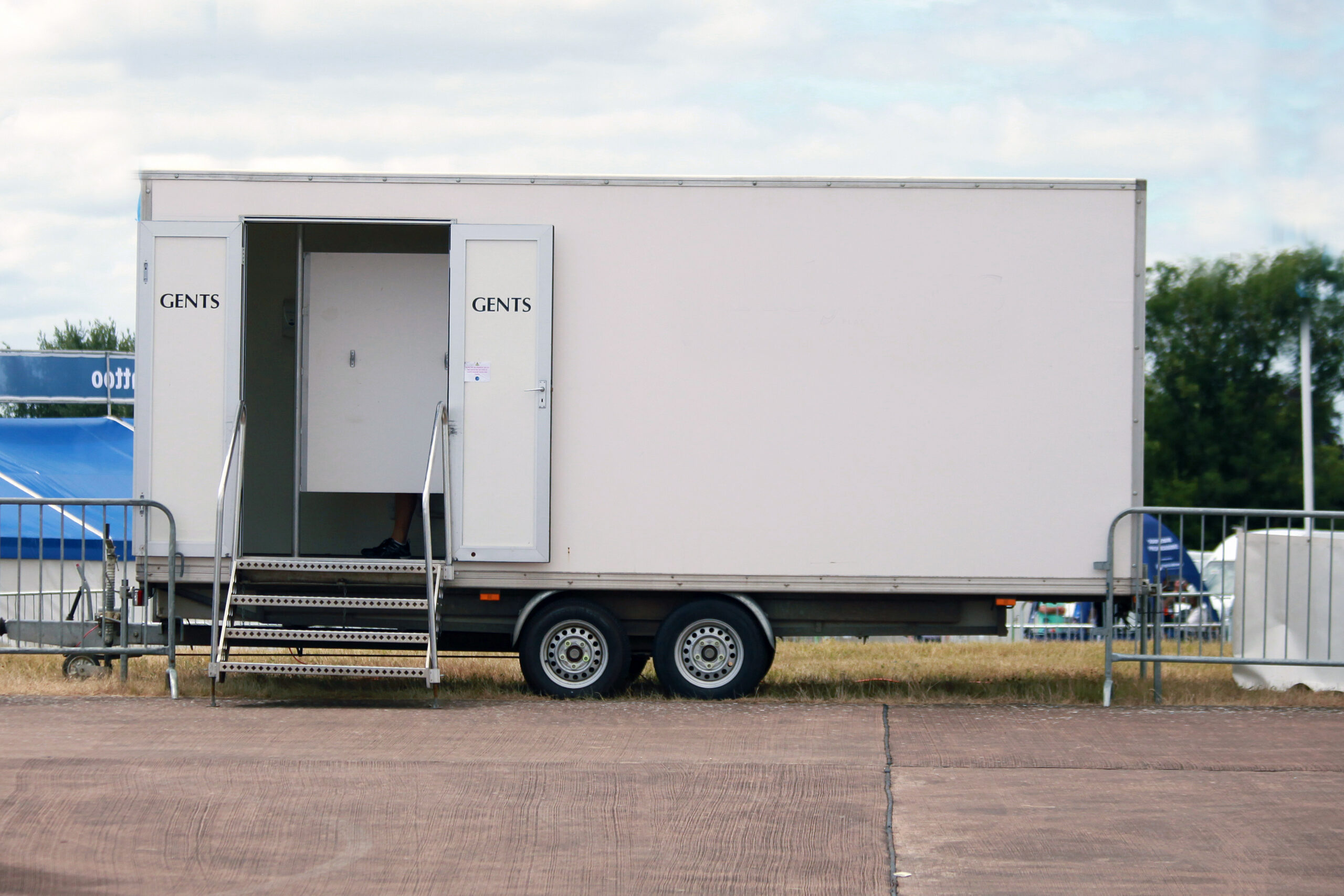 A white portable restroom trailer labeled "GENTS" with stairs leading to the entrance, parked outdoors on a paved surface, with grass, barriers, and tents visible in the background.