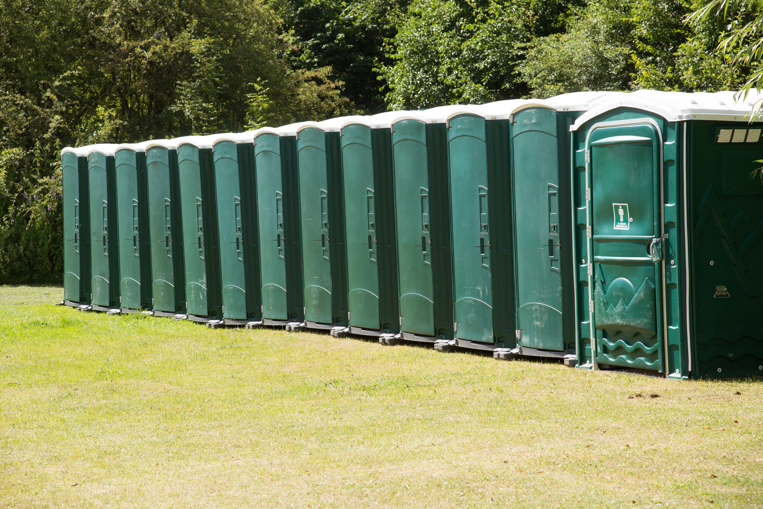 A row of green portable toilets lined up on grass in an outdoor setting, surrounded by trees and greenery.