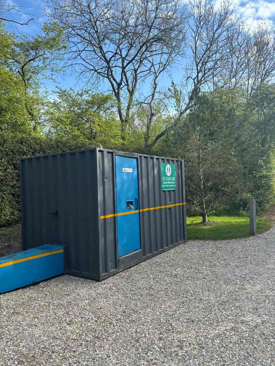 A dark gray metal storage container with a blue door and yellow stripe sits on a gravel path, surrounded by trees and greenery under a clear sky.