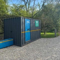 A dark gray portable container with a bright blue door and yellow stripe sits on gravel near trees and greenery. A green sign on the container reads "Site Lock Up.