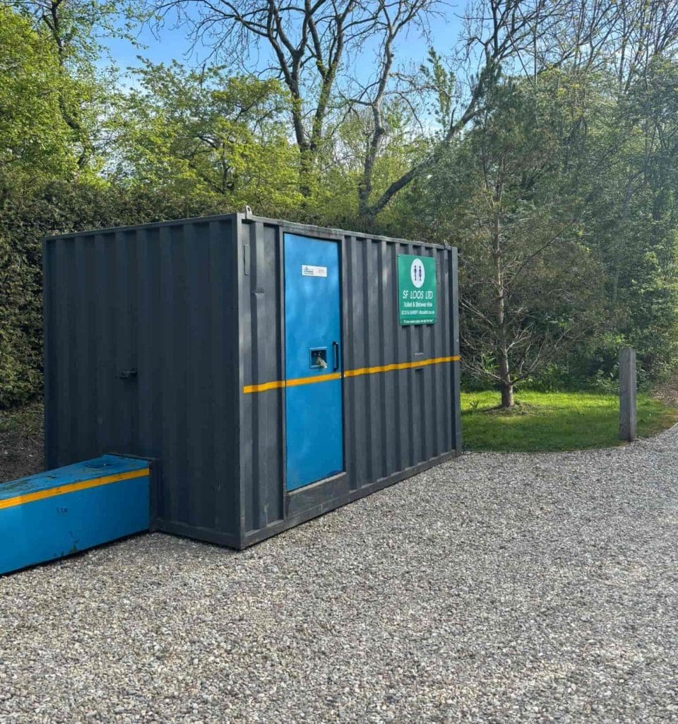 A dark gray portable container with a bright blue door and yellow stripe sits on gravel near trees and greenery. A green sign on the container reads "Site Lock Up.