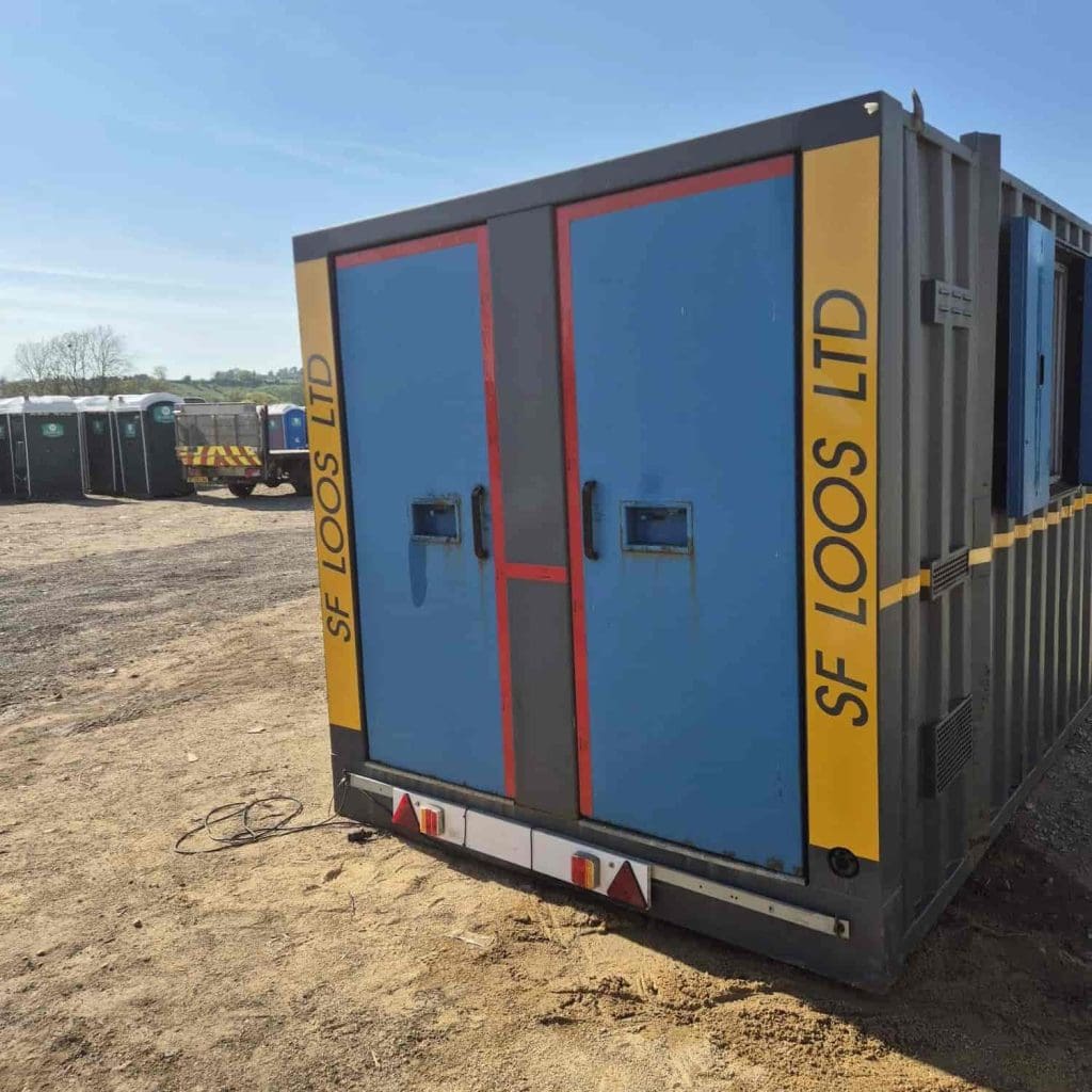 A large gray and blue portable toilet unit with "SF LOOS LTD" written on yellow stripes stands on a dirt lot; smaller portable toilets are visible in the background.